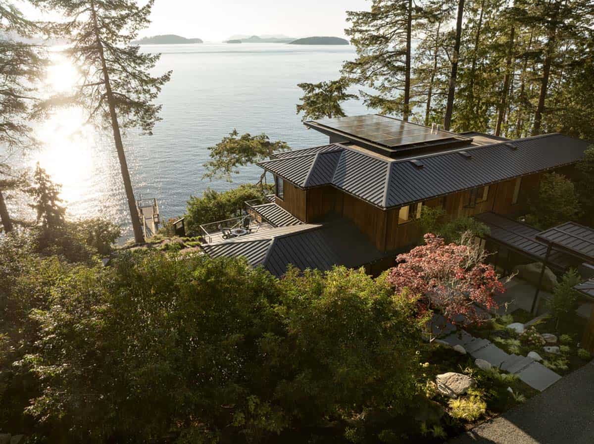 Aerial view of modern waterfront home with dark metal roof and solar panels nestled among Pacific Northwest trees