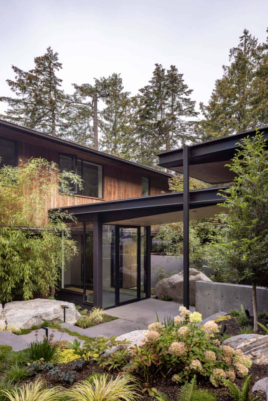 Black steel canopy entry with floor-to-ceiling glass doors surrounded by hydrangeas, ferns, and native boulders