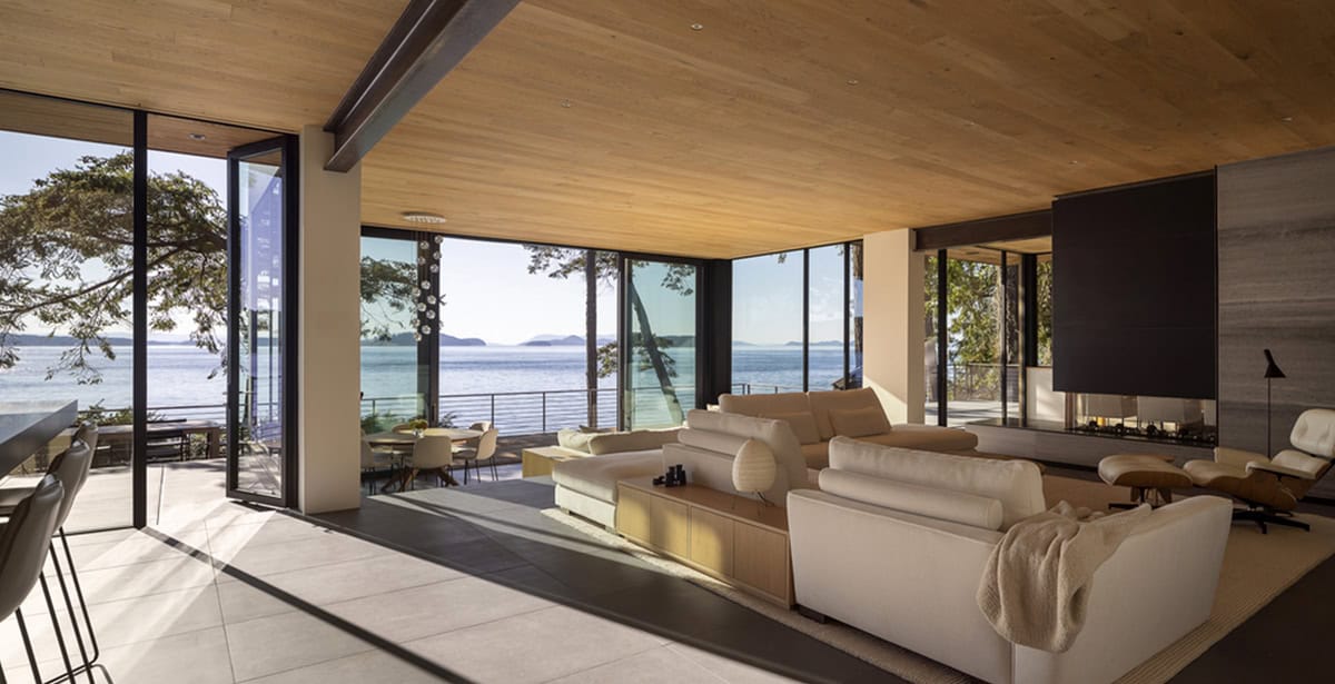 Open-plan living room with wood ceiling, floor-to-ceiling windows, cream sofas, and sweeping island water views