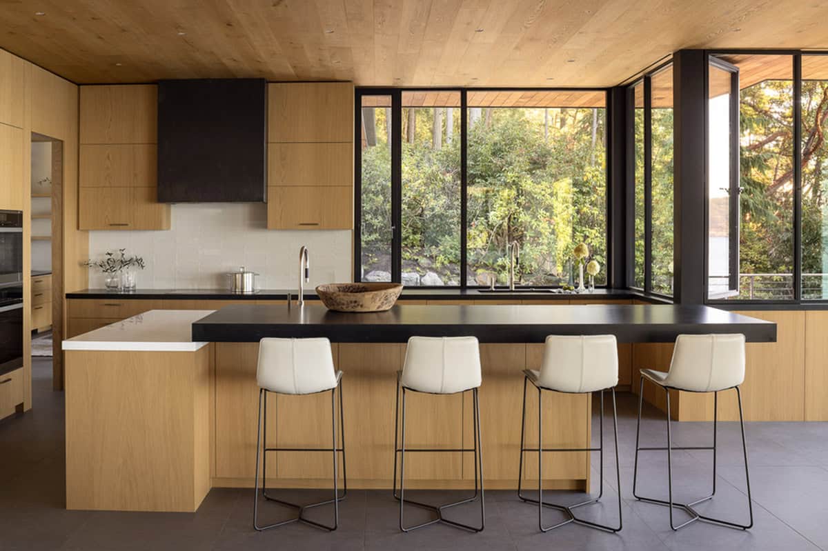 Modern kitchen with light oak cabinetry, black island countertop, cream barstools, and garden-view windows