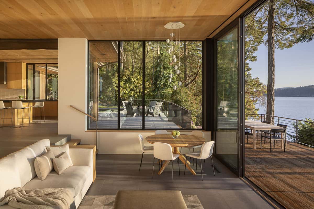 Dining nook with round wood table beside floor-to-ceiling windows and open sliding doors to waterfront deck