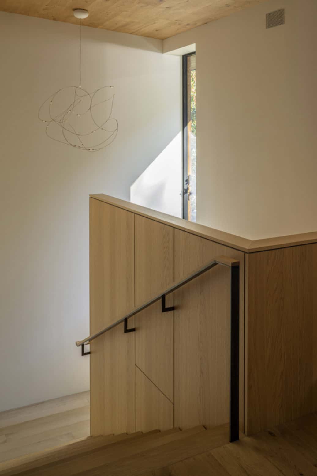 Upper stair landing with oak paneling, metal handrail, sculptural wire pendant light, and clerestory window