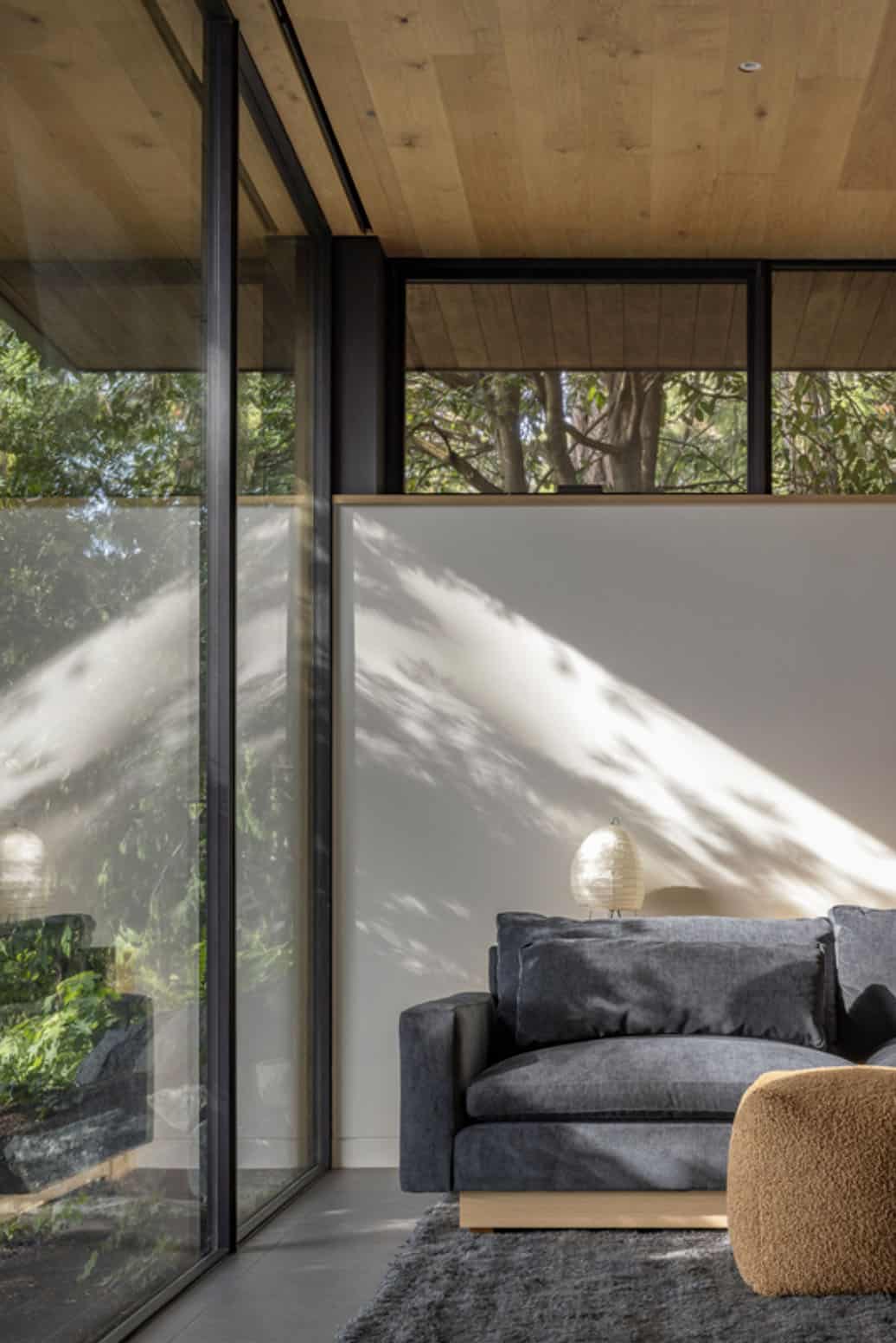 Bedroom sitting area with charcoal sofa, dappled tree shadow on white wall, and floor-to-ceiling garden windows