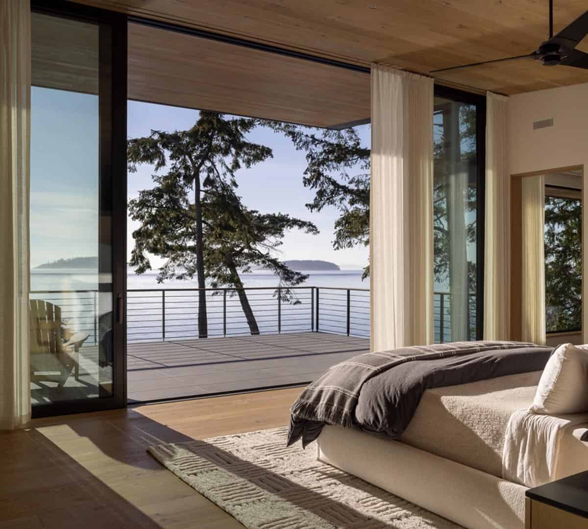 Primary bedroom with low platform bed, sheer curtains, wood floors, and open glass doors to water-view balcony