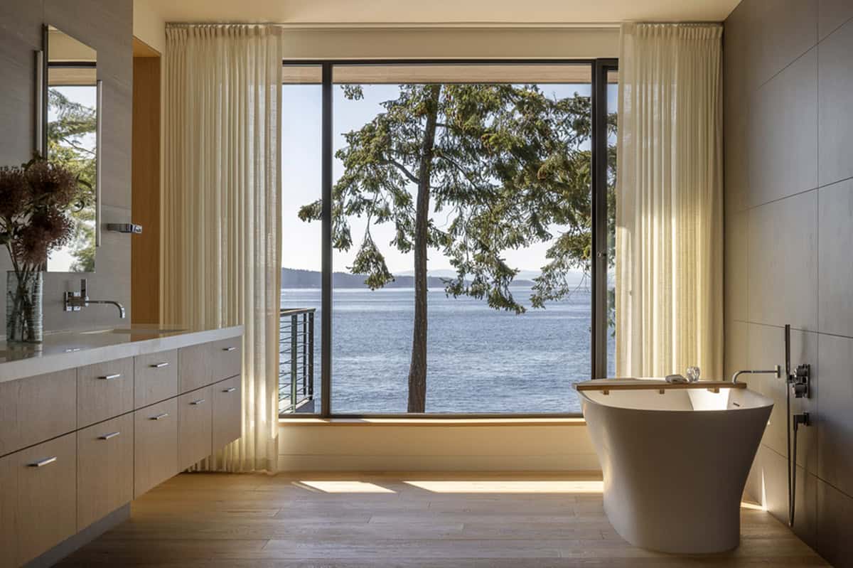 Freestanding soaking tub beside large picture window with sheer curtains framing a waterfront tree view