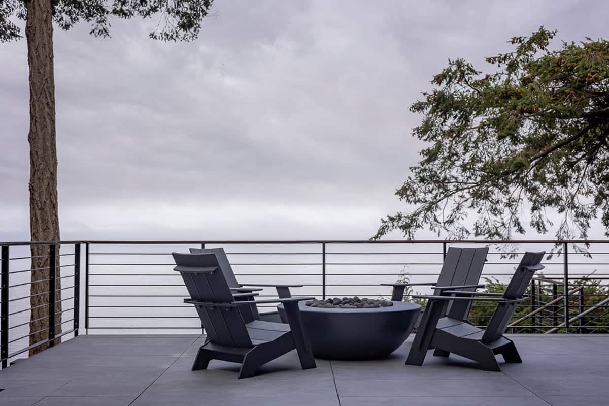 Modern waterfront deck with dark Adirondack chairs around a round fire bowl under overcast skies