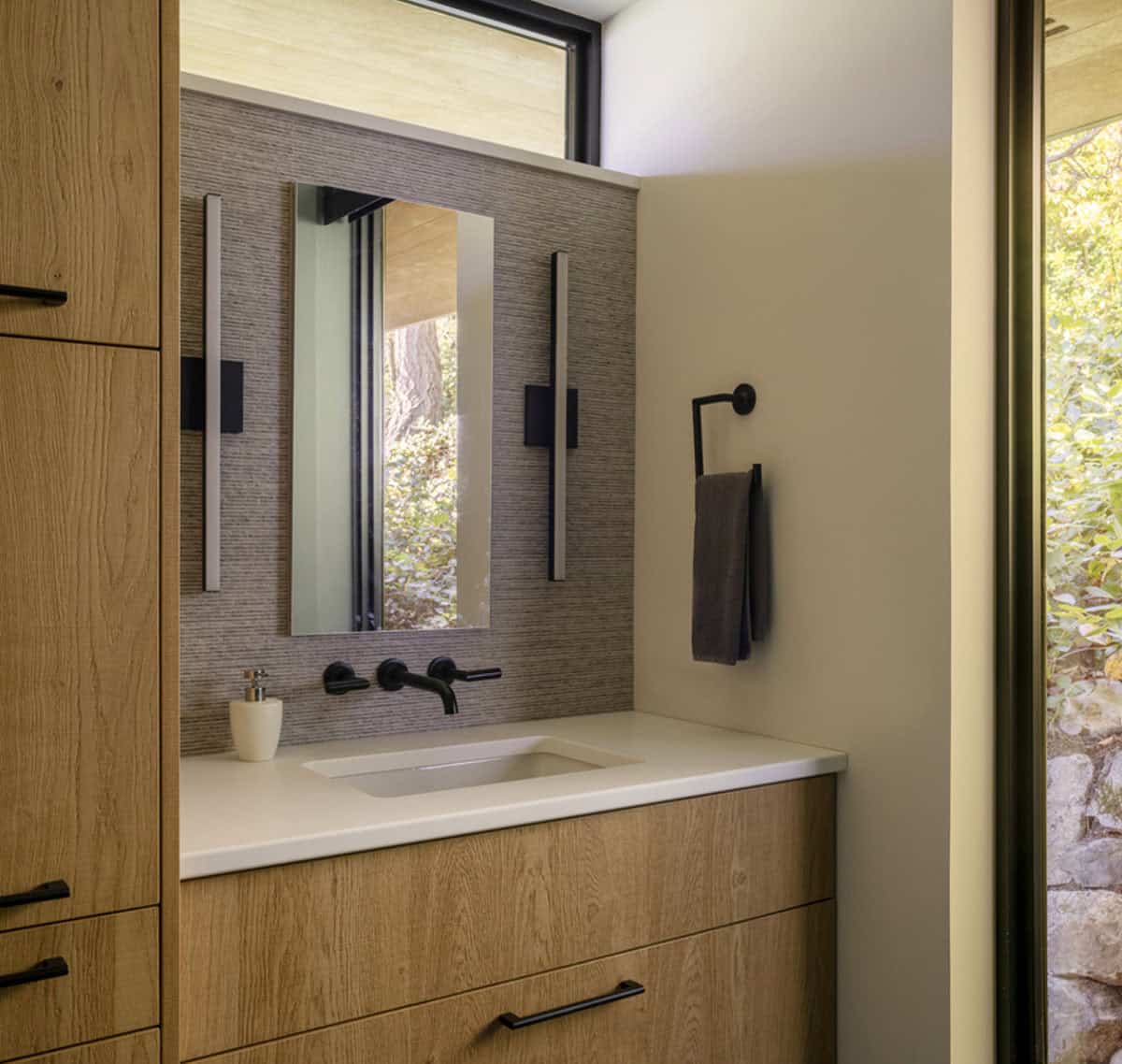 Oak bathroom vanity with matte black fixtures, textured tile backsplash, and garden views through clerestory window