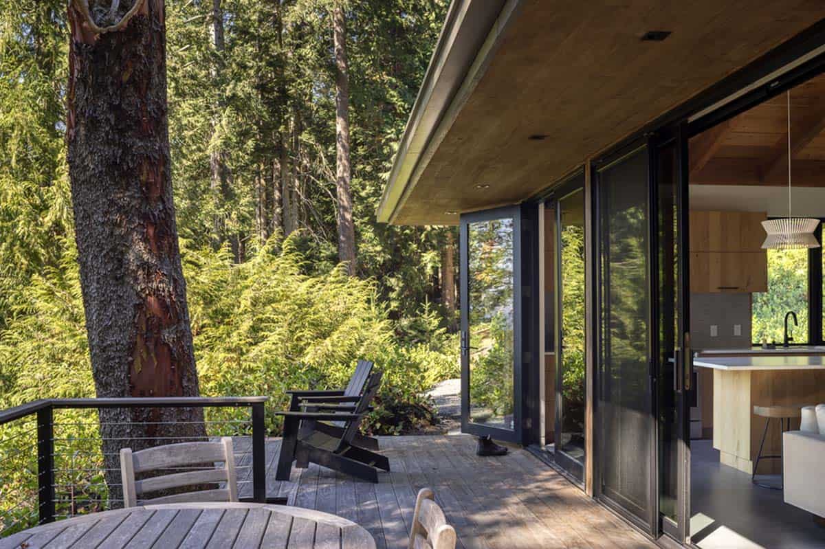 Forested deck off guest cottage with Adirondack chairs, wood dining table, and open sliding doors to kitchen