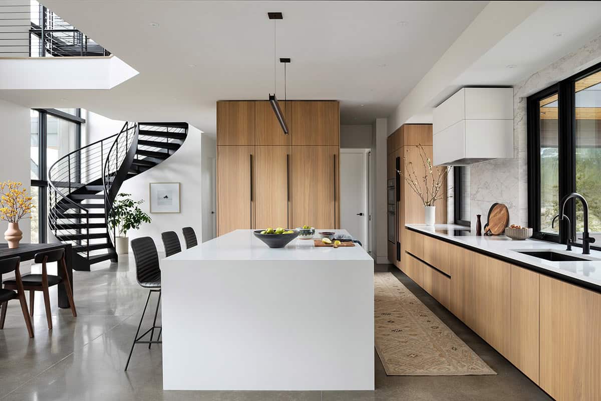Modern kitchen with white waterfall island, warm wood cabinetry, marble backsplash, and black spiral staircase
