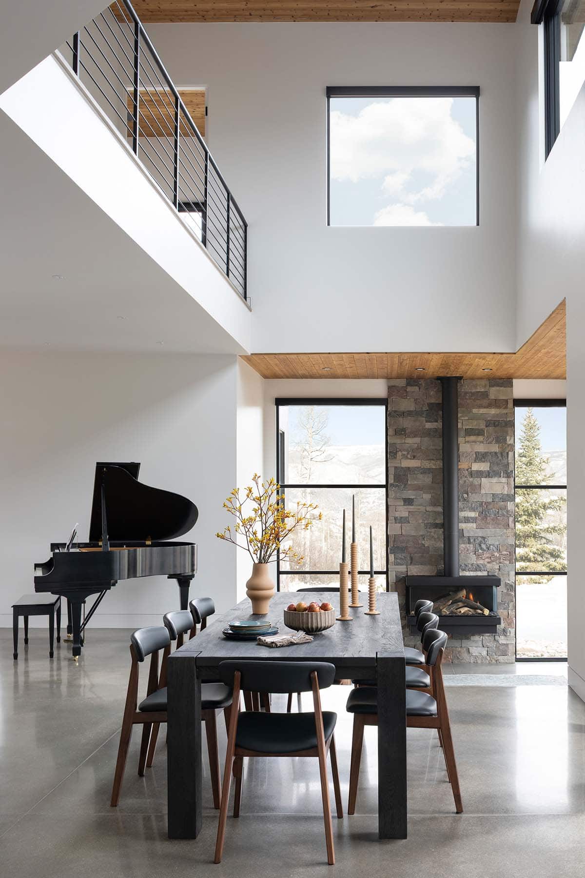 Dining room with dark wood table, mid-century chairs, grand piano, stone fireplace, and double-height windows