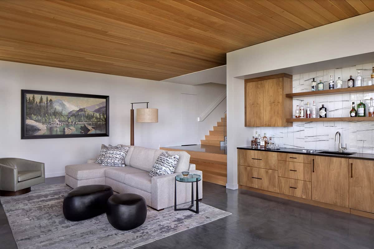 Lower level lounge with wood ceiling, built-in wet bar with open shelving, sectional sofa, and a framed mountain landscape painting
