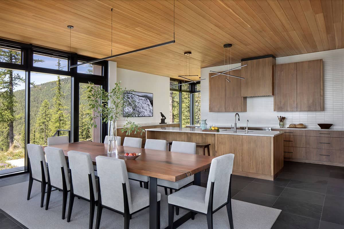 Open-plan dining and kitchen with a live-edge walnut table, walnut cabinetry, white tile backsplash, and floor-to-ceiling windows overlooking a forested mountainside