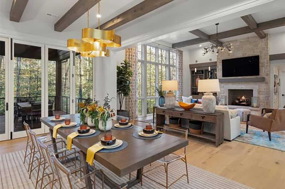 Open dining area with gold chandelier looking toward stone fireplace living room and wooded views