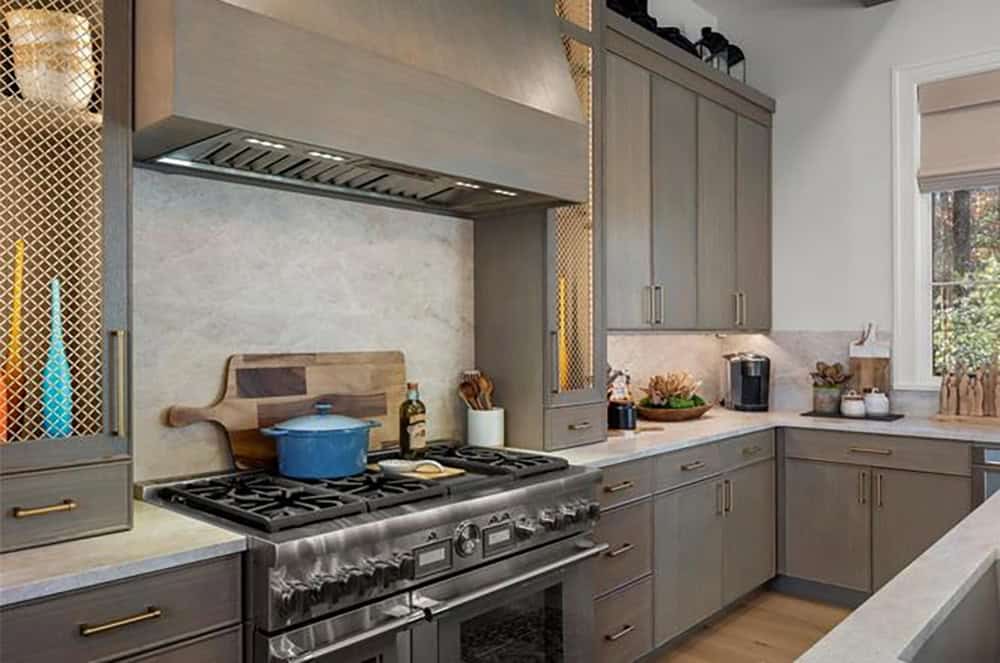 Close-up of professional gas range with custom hood, gray shaker cabinets, stone backsplash, and brass hardware
