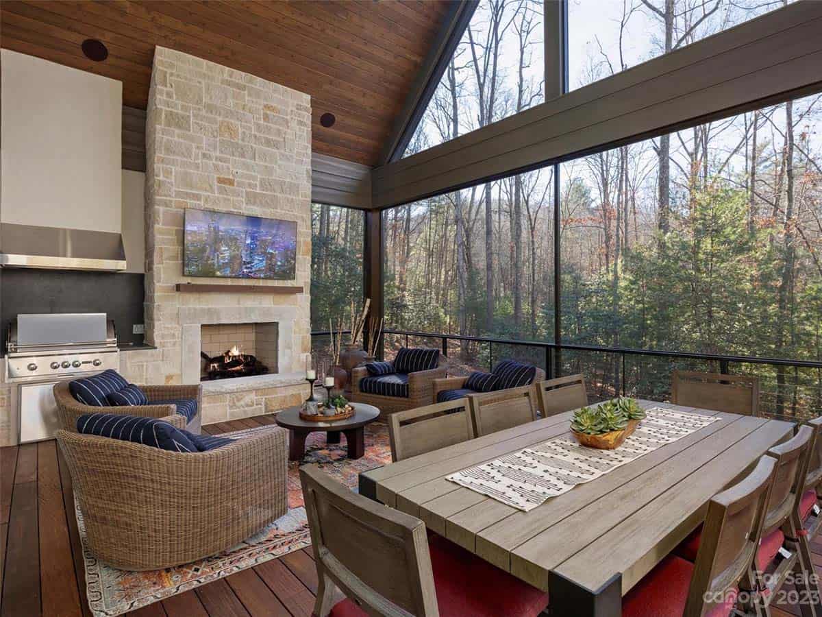 Screened porch with vaulted wood ceiling, stone fireplace, outdoor kitchen, wicker seating, and wooded views