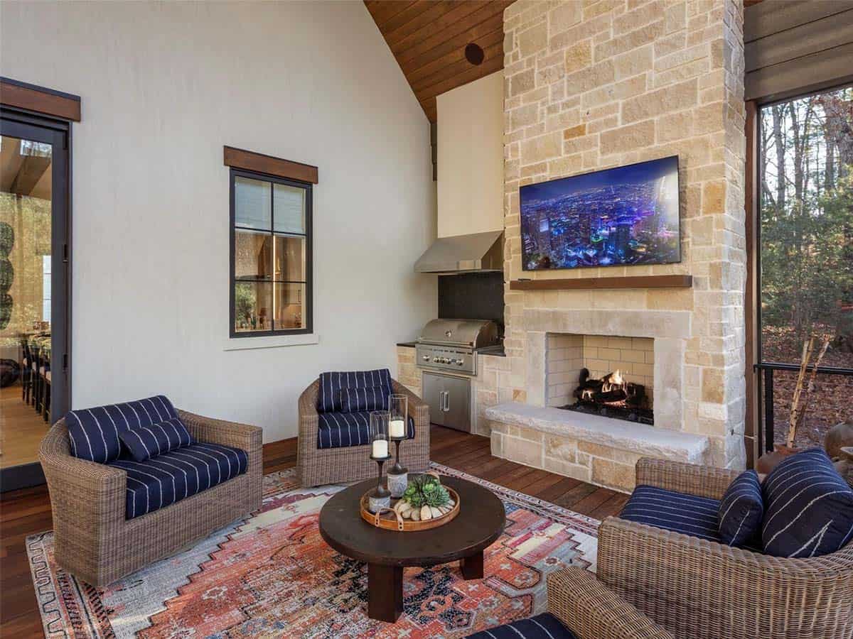 Covered outdoor living space with stone fireplace, TV, outdoor kitchen, wicker seating, and screened wooded views