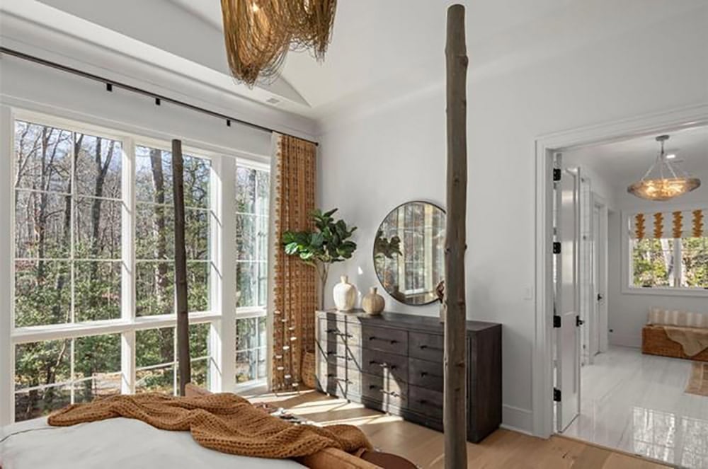 Primary bedroom with natural wood four-poster bed frame, large windows overlooking woods, and woven pendant light