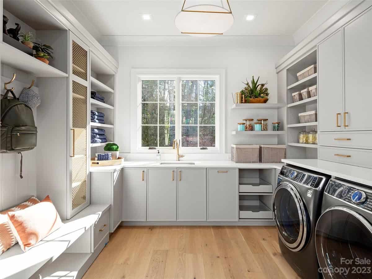 Spacious laundry and mudroom with light gray cabinets, built-in shelving, brass hardware, and front-load washer/dryer