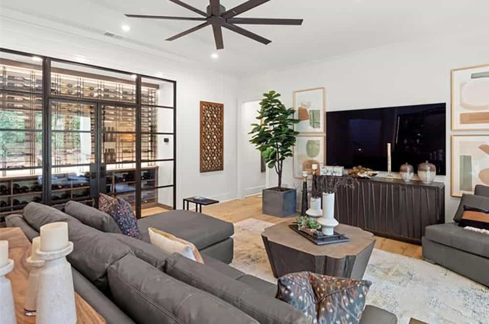 Lower-level media room with gray sectional, large TV, and glass-enclosed wine cellar visible beyond