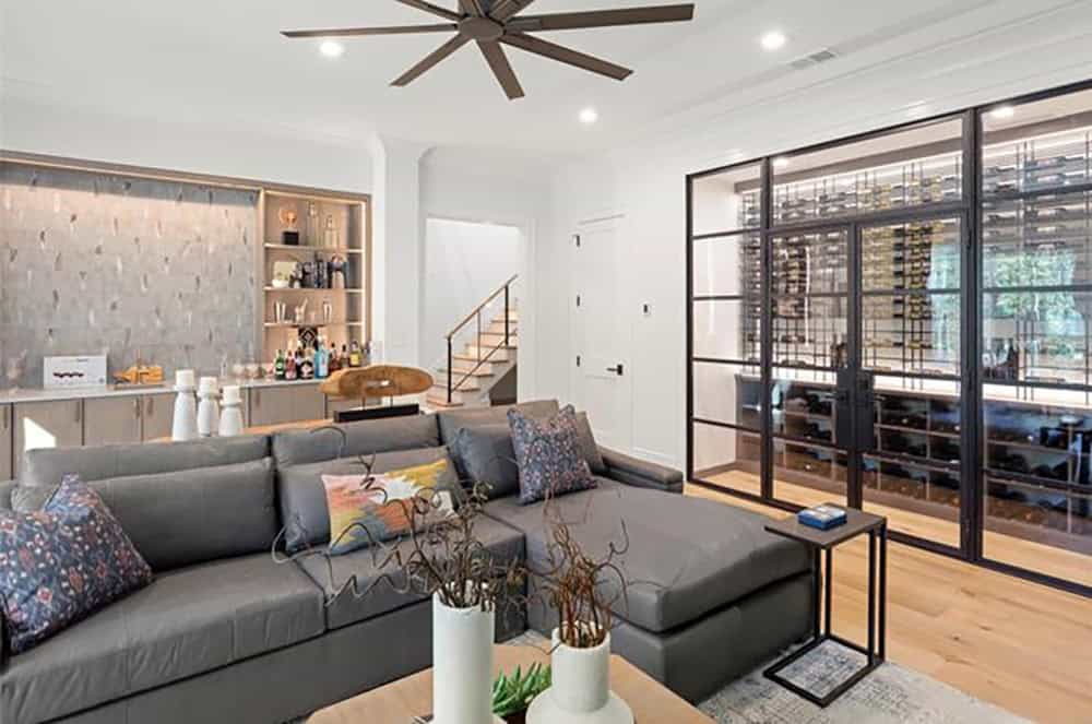 Lower-level lounge with gray sectional, wet bar with textured backsplash, and glass-walled wine cellar