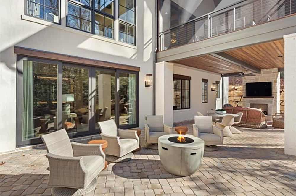 Lower patio with brick pavers, round gas fire pit, wicker seating, and covered outdoor living area beyond