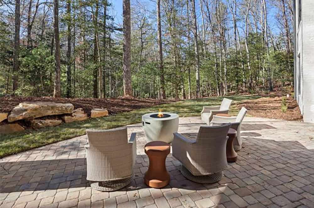 Brick paver patio with round gas fire pit, wicker swivel chairs, and wooded backyard views