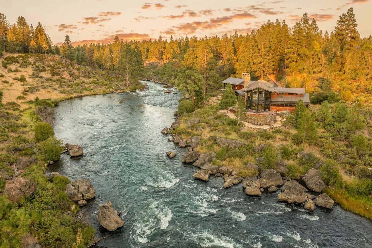 Aerial view of a modern riverside retreat in Oregon at sunset, surrounded by pine trees and rocky rapids