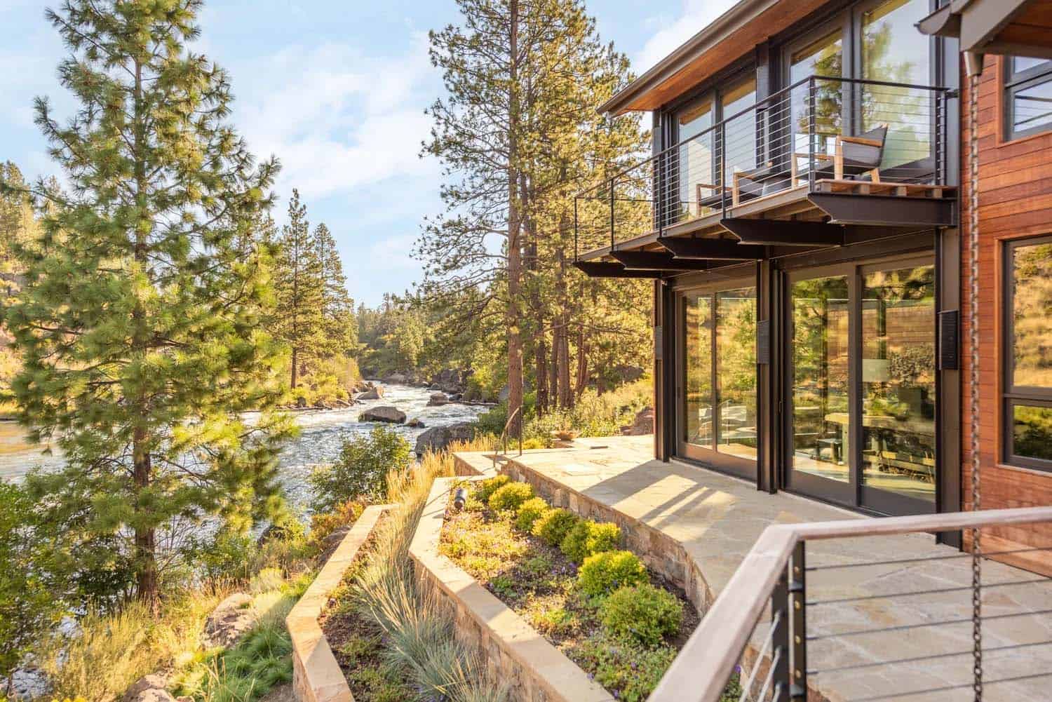 Riverside terrace of a modern mountain home with cantilevered balcony overlooking pine forest and rapids
