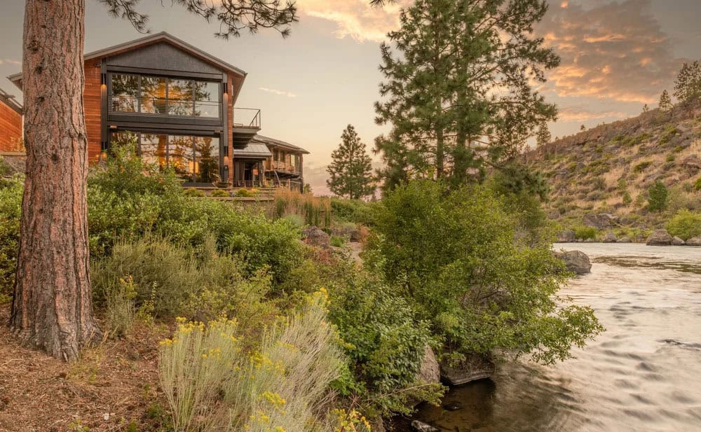 Side view of a multi-story glass and cedar home perched above a river at dusk, flanked by pine trees