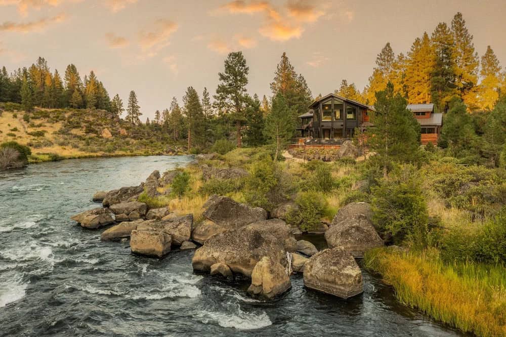 Distant view of a modern wood-and-glass river home across rocky whitewater rapids at dusk