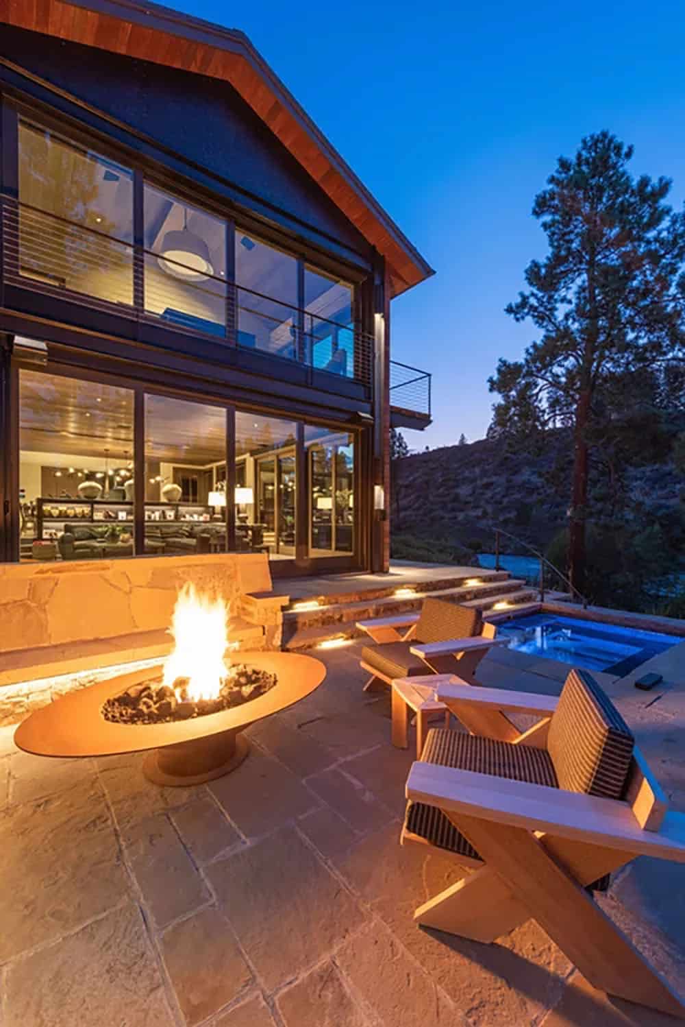 Outdoor patio at night with a glowing fire bowl, lounge chairs, and illuminated hot tub beside a glass-walled home