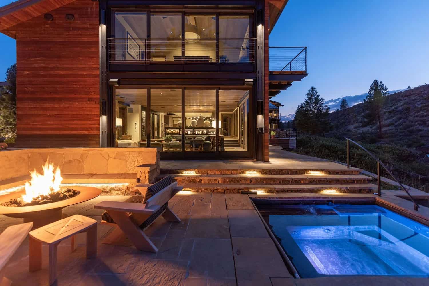 Evening view of a cedar and glass home's back patio with lit fire bowl, hot tub, and stepped stone terrace