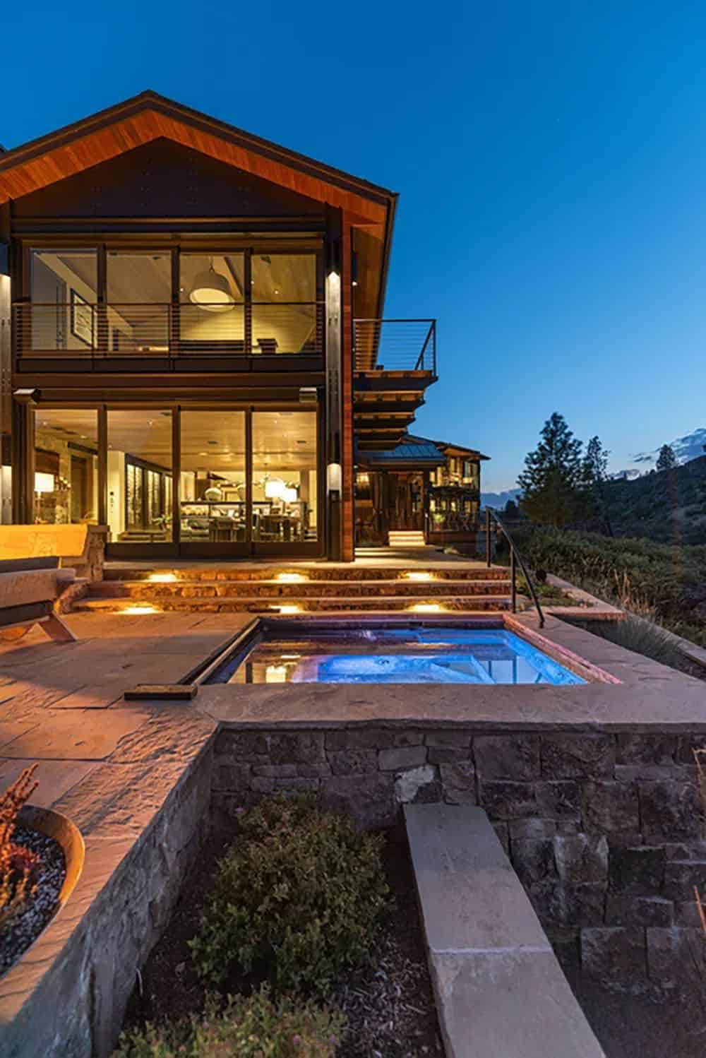 Night view of a two-story glass home exterior with glowing hot tub, lit stone steps, and mountain backdrop
