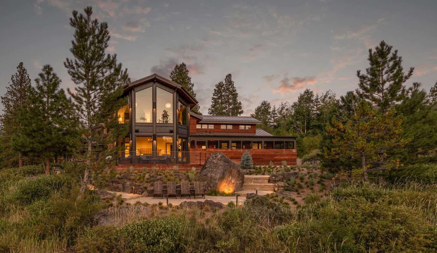 Twilight exterior of a modern glass-gabled home nestled among pines, with lit Adirondack chairs and boulder landscape