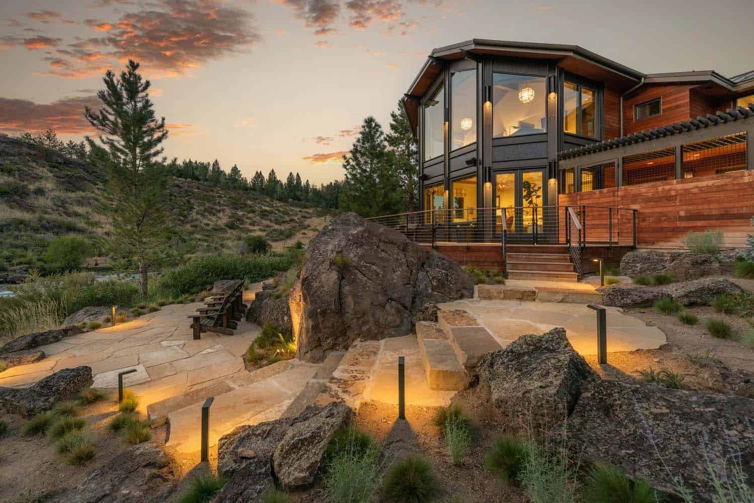 Illuminated flagstone pathway winding through boulders toward a glowing glass tower home at sunset