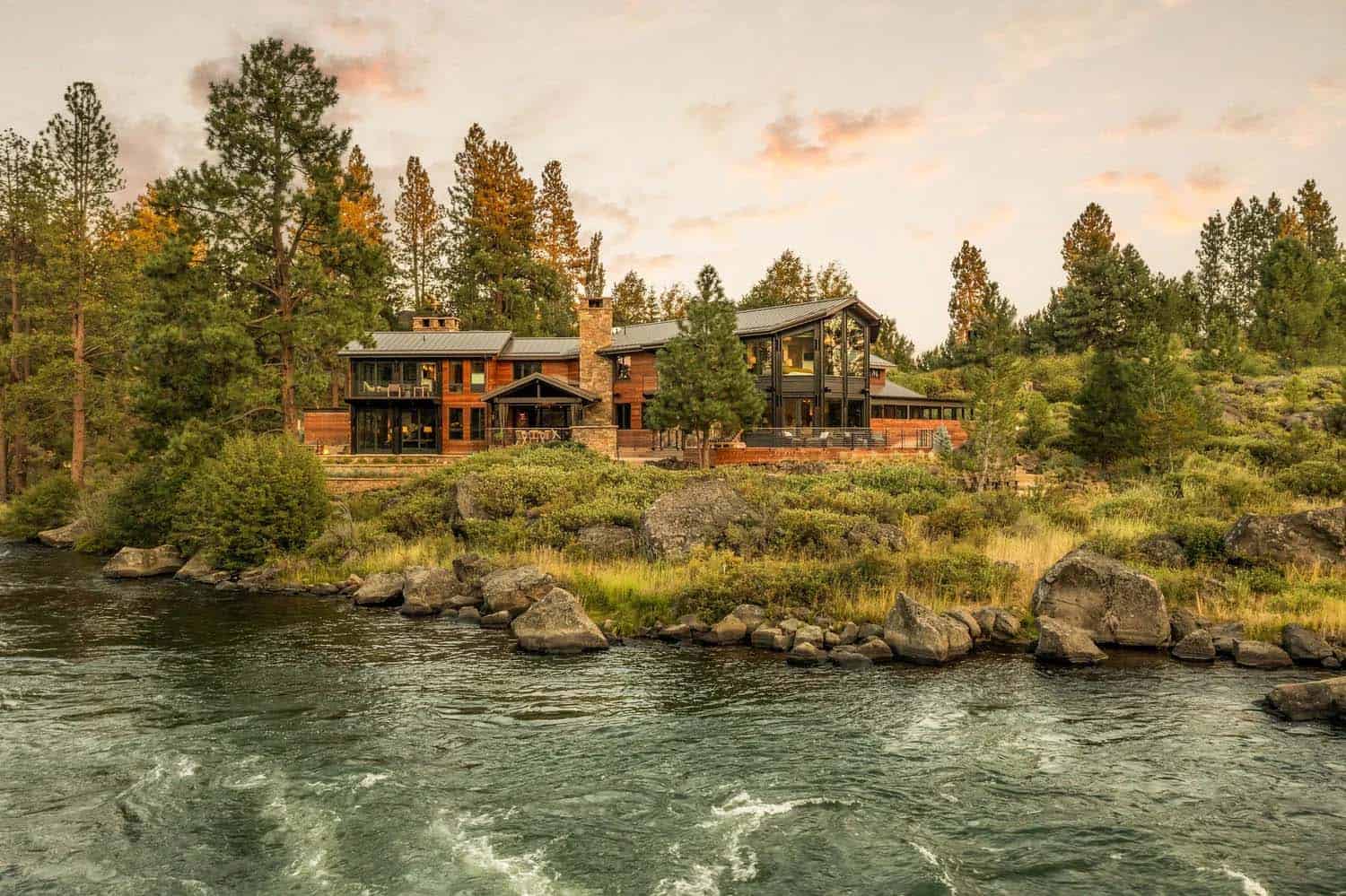 River-level view of two connected modern cedar homes on a forested Oregon riverbank at sunset