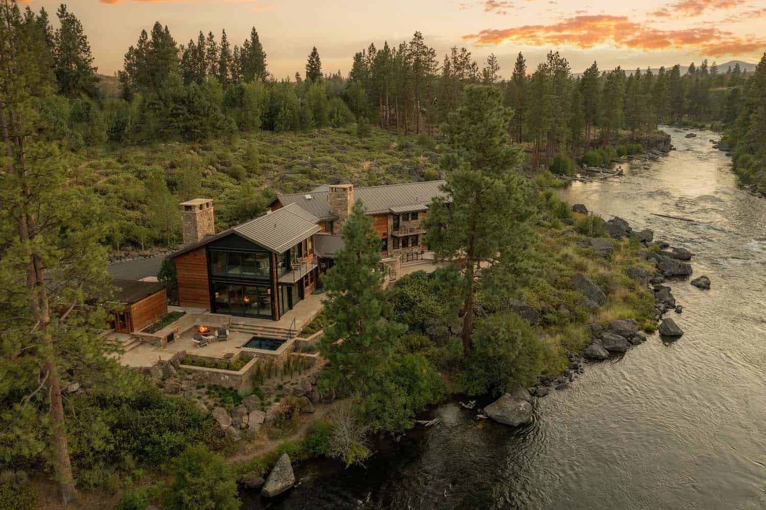 Bird's-eye view of a cedar and glass river home with fire pit and hot tub terrace nestled among pines