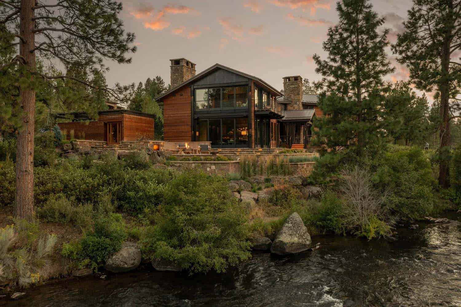 Riverfront view of a cedar home with stone chimney and terraced landscaping glowing at dusk