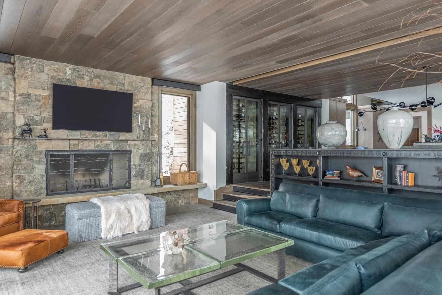 Living room with stone fireplace, wall-mounted TV, teal leather sectional, and glass wine cellar beyond