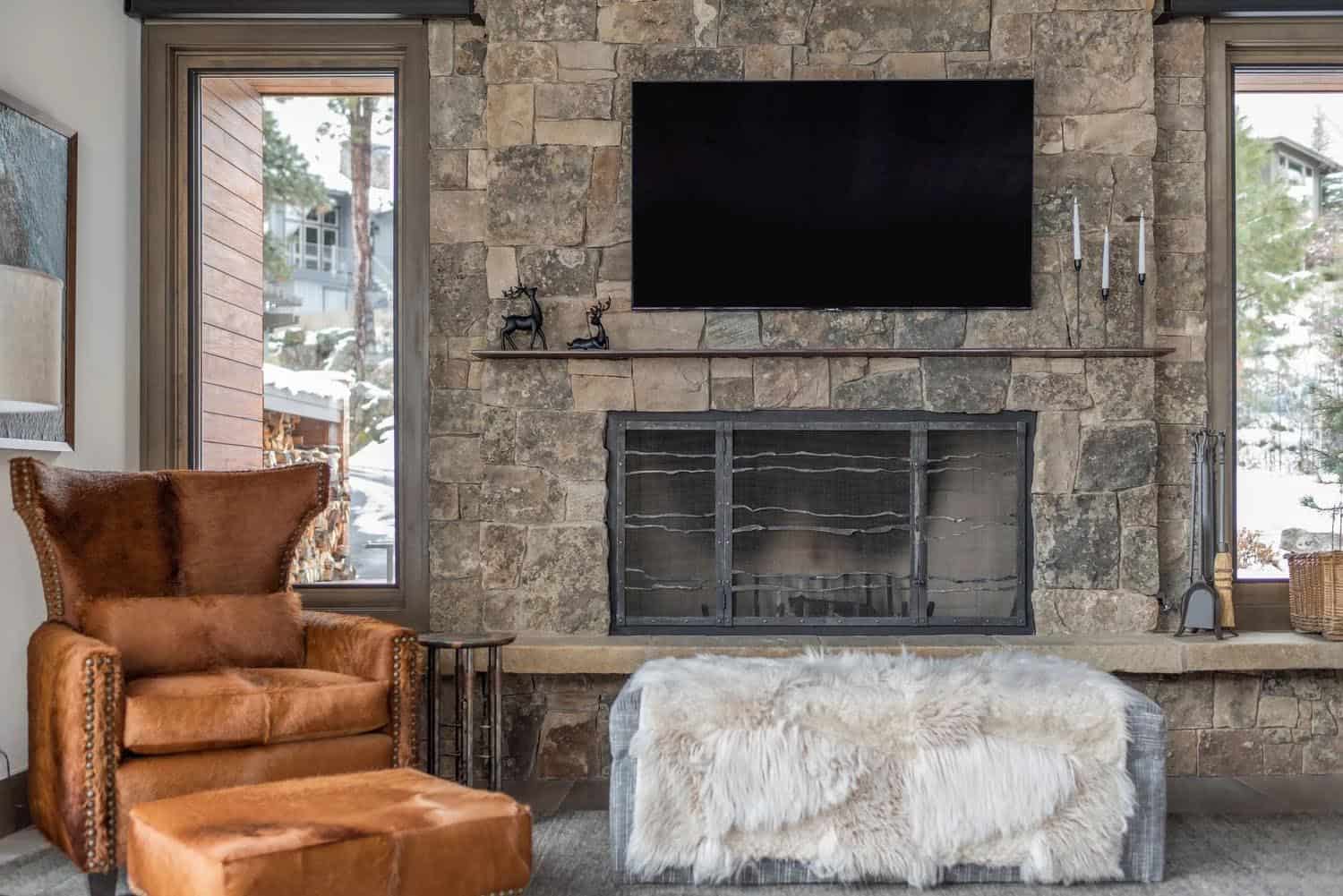 Close-up of a rustic stone fireplace with mounted TV, leather wingback chair, and sheepskin ottoman