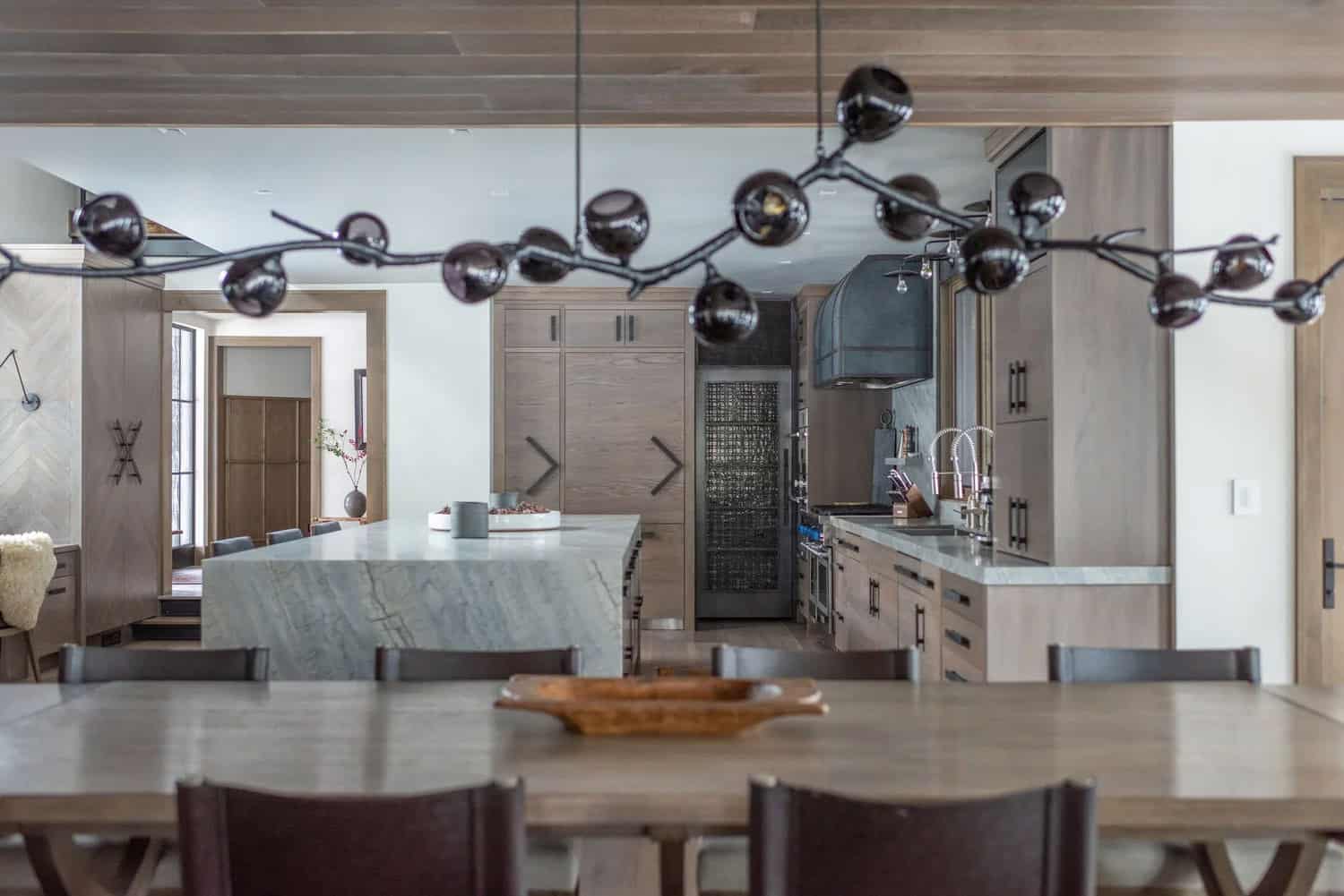 Open kitchen viewed through a branch chandelier, with marble island, custom cabinetry, and range hood beyond
