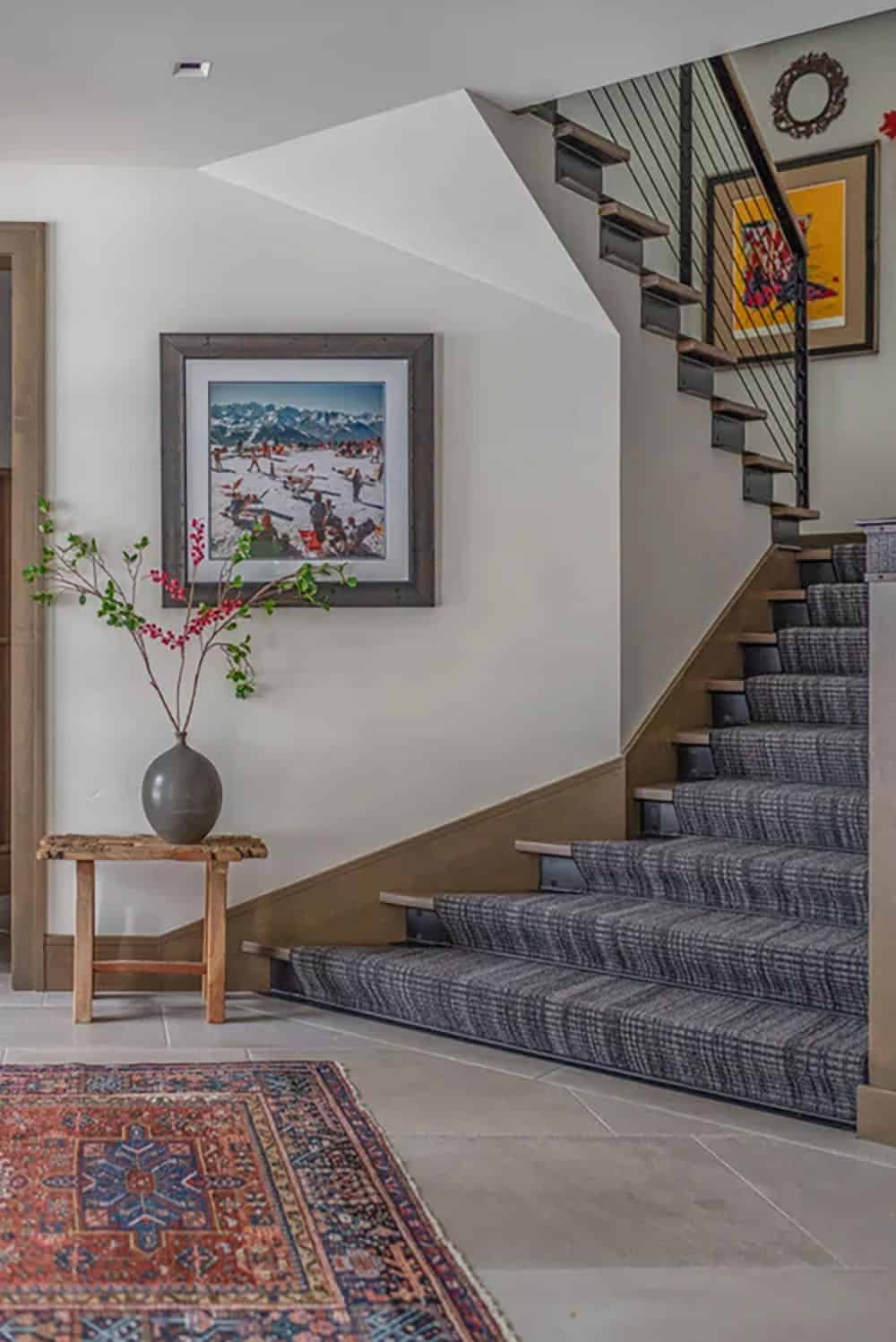 Entryway staircase with carpeted treads, cable railing, framed ski art, and Persian rug on tile floor