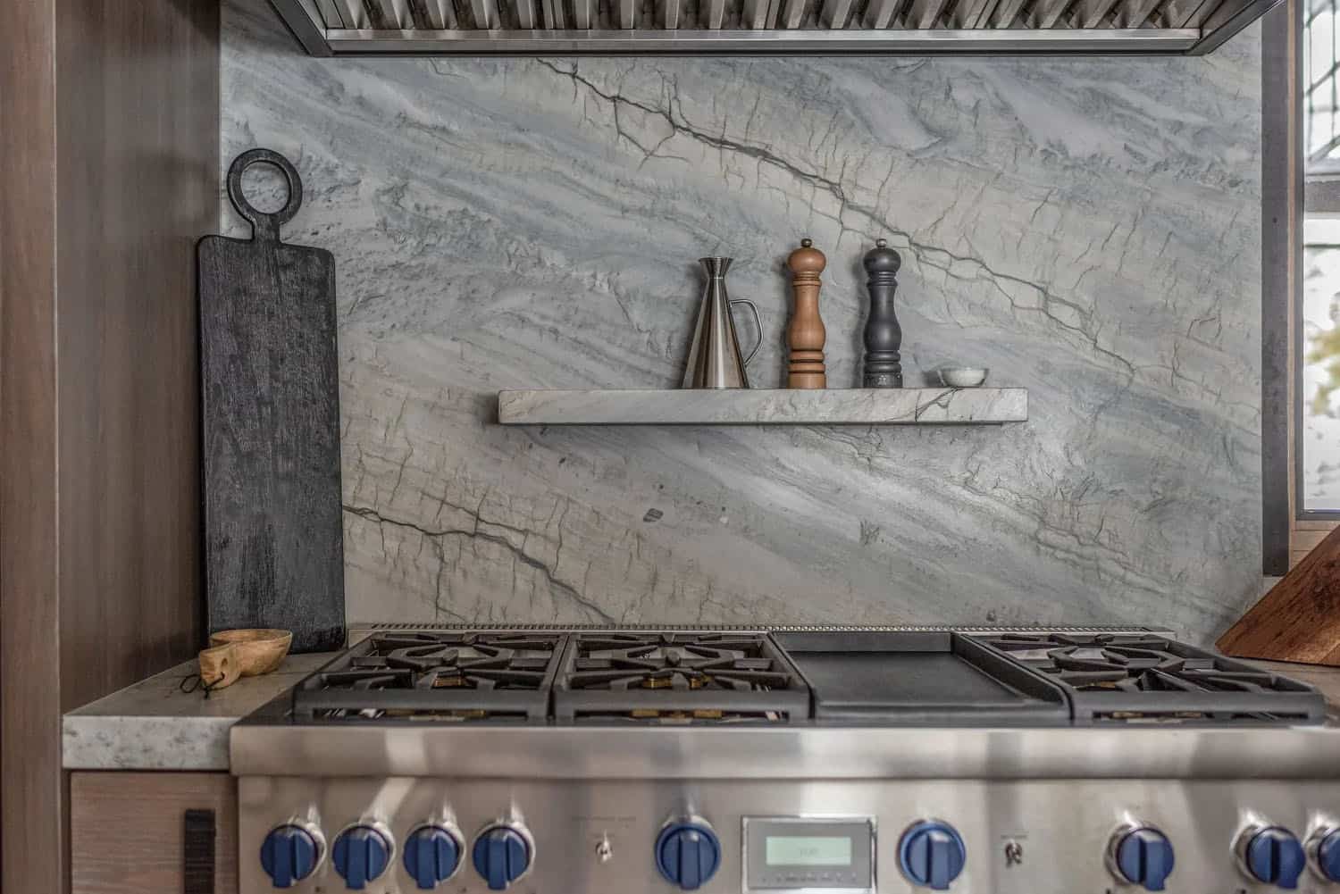 Close-up of professional kitchen range with dramatic veined marble slab backsplash and floating stone shelf