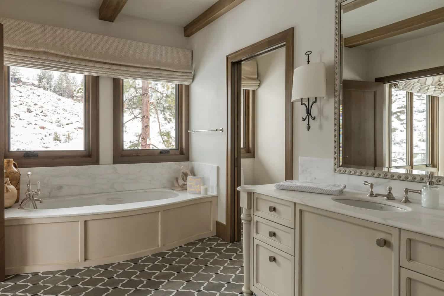 Bright bathroom with marble soaking tub, geometric tile floor, wood beam ceiling, and snowy forest views