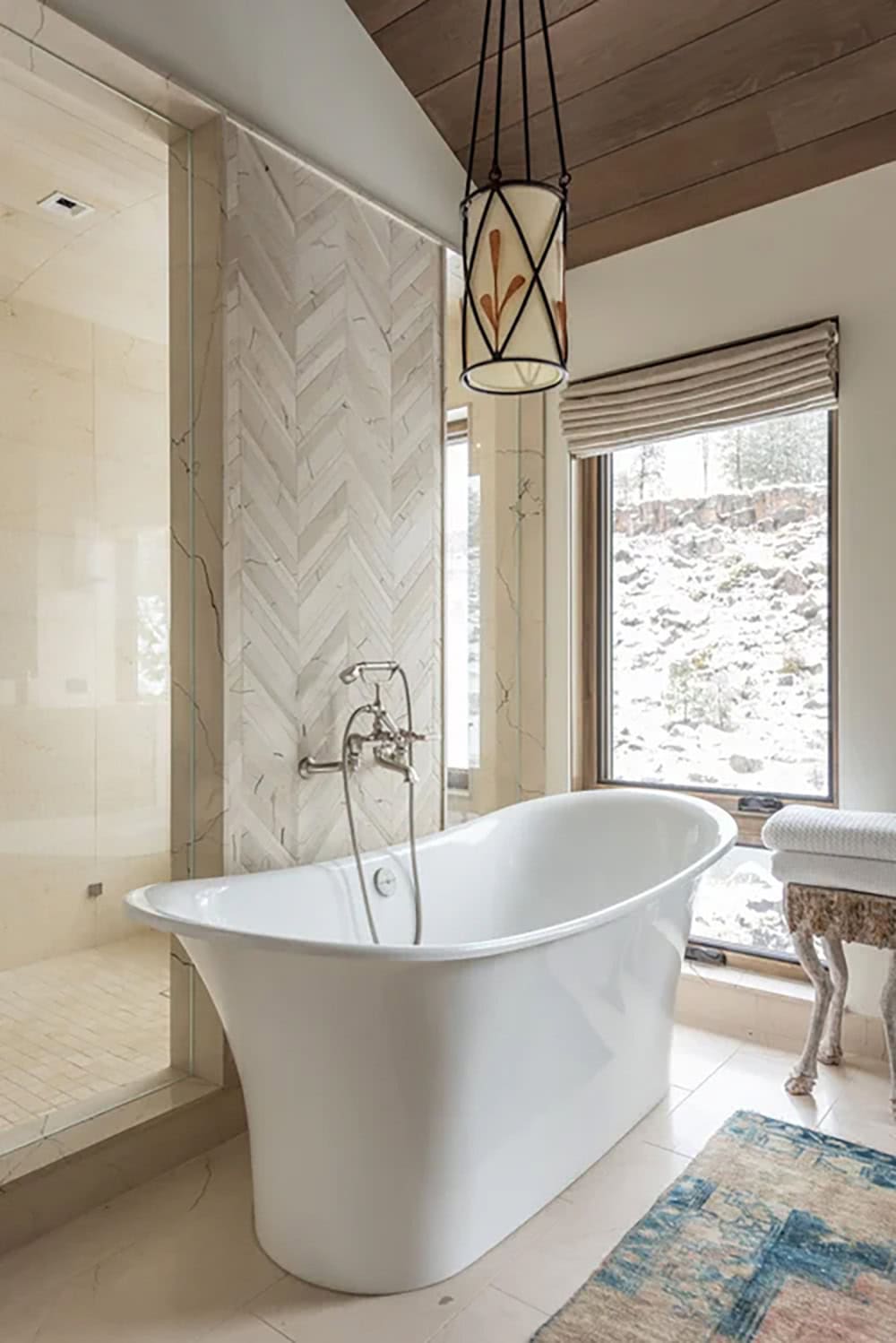 Freestanding soaking tub beside herringbone marble feature wall with pendant light and snowy window views