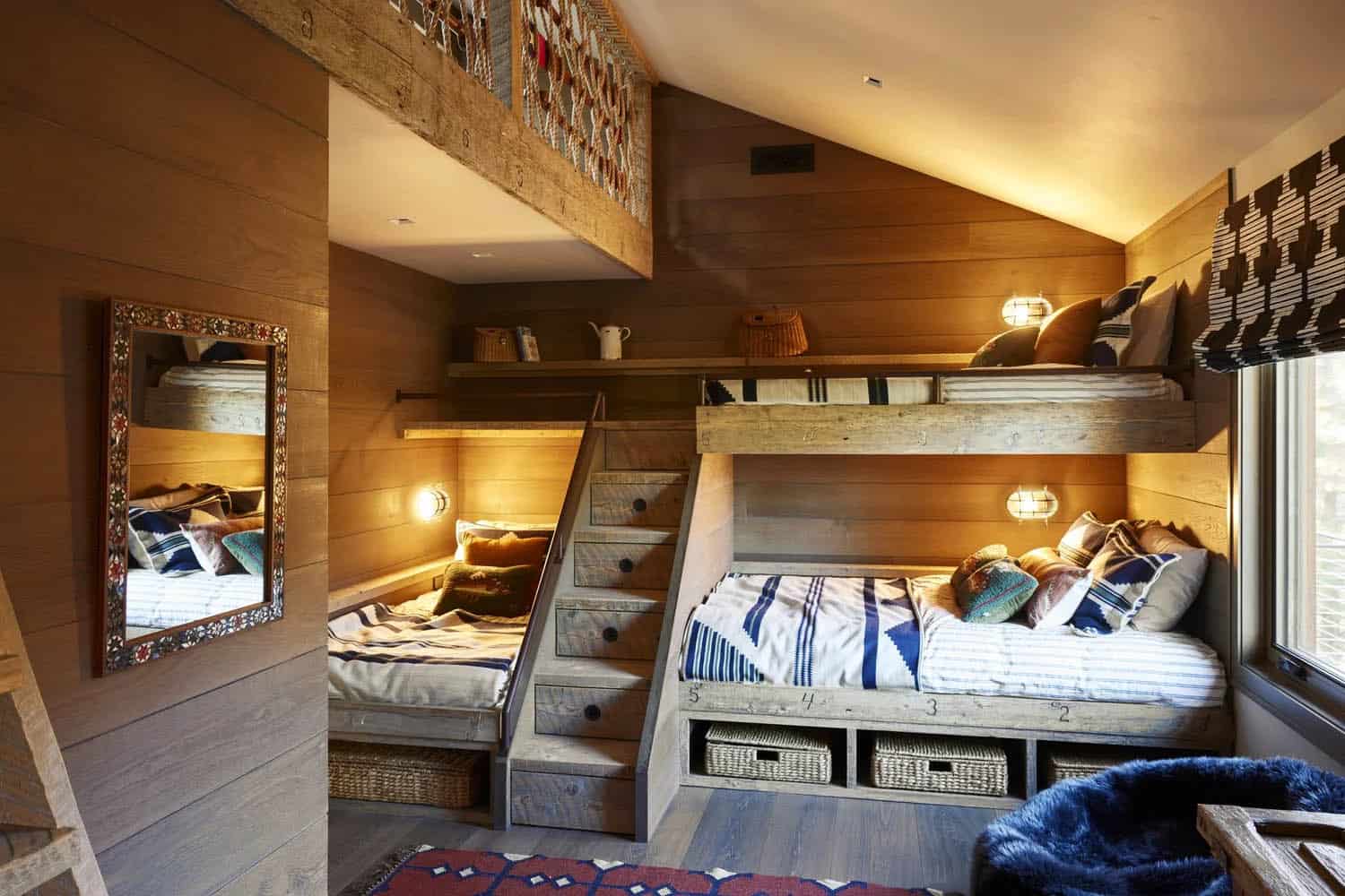 Rustic bunk room with built-in stacked wood bunks, stair drawers, warm sconce lighting, and mezzanine loft
