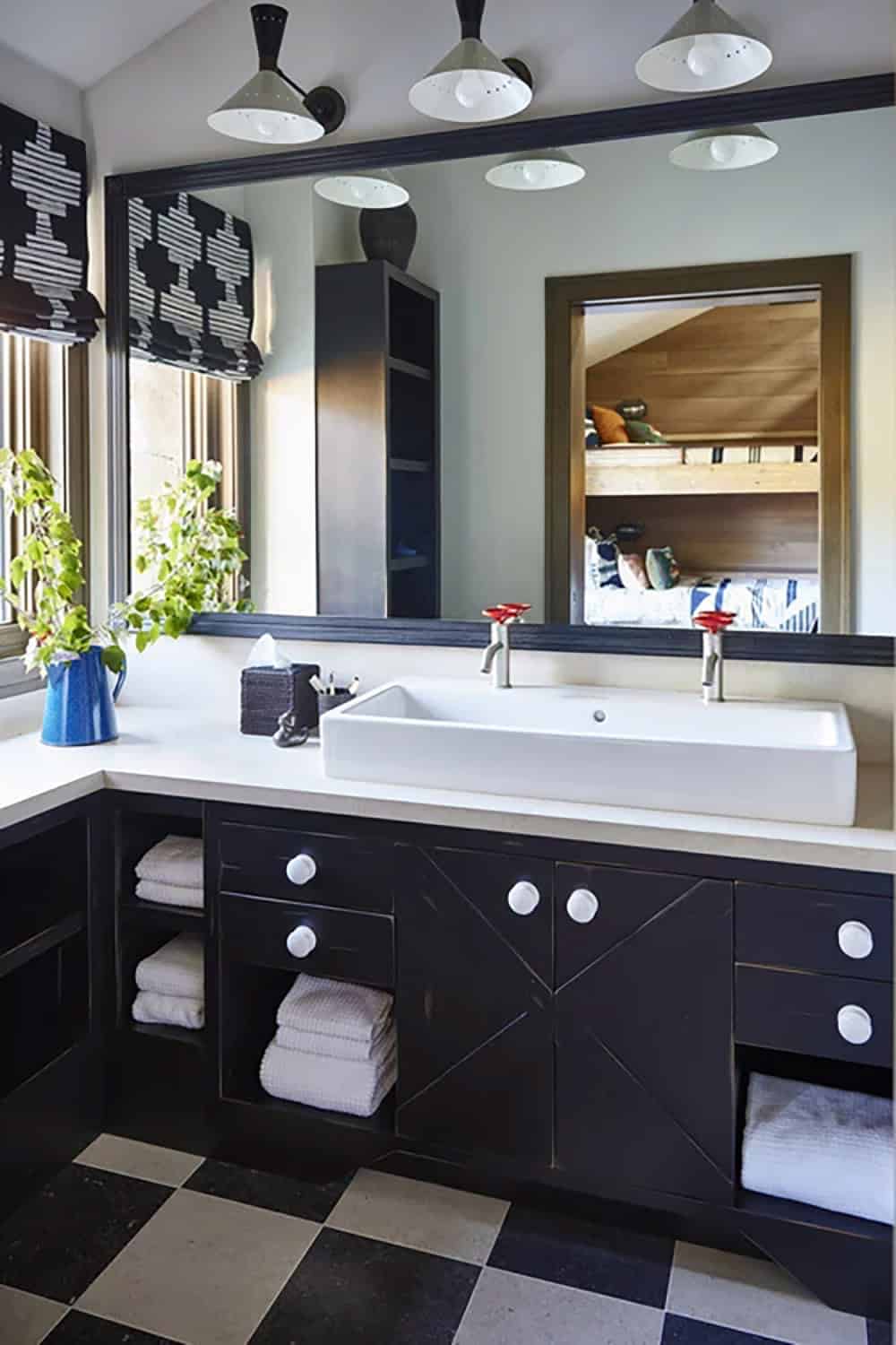 Kids' bathroom with black cabinetry, trough sink, red faucets, checkerboard tile floor, and mirror reflecting bunk room