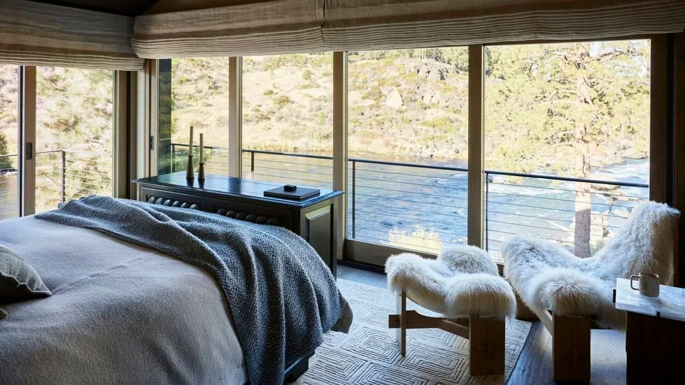 Primary bedroom with floor-to-ceiling windows overlooking the river, sheepskin lounge chair, and gray bedding