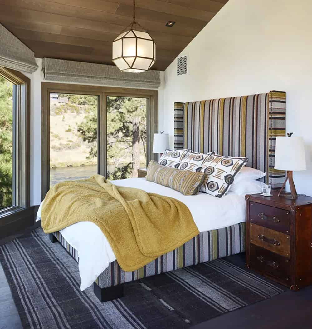 Guest bedroom with striped upholstered headboard, mustard throw, geometric pendant light, and river views