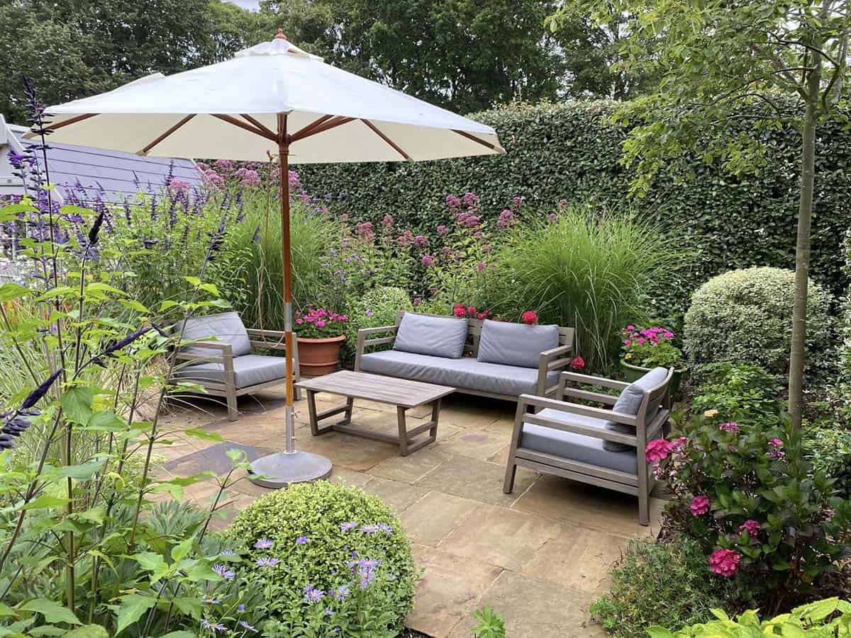 Lush cottage garden patio with grey outdoor sofa set surrounded by hedges and flowering perennials